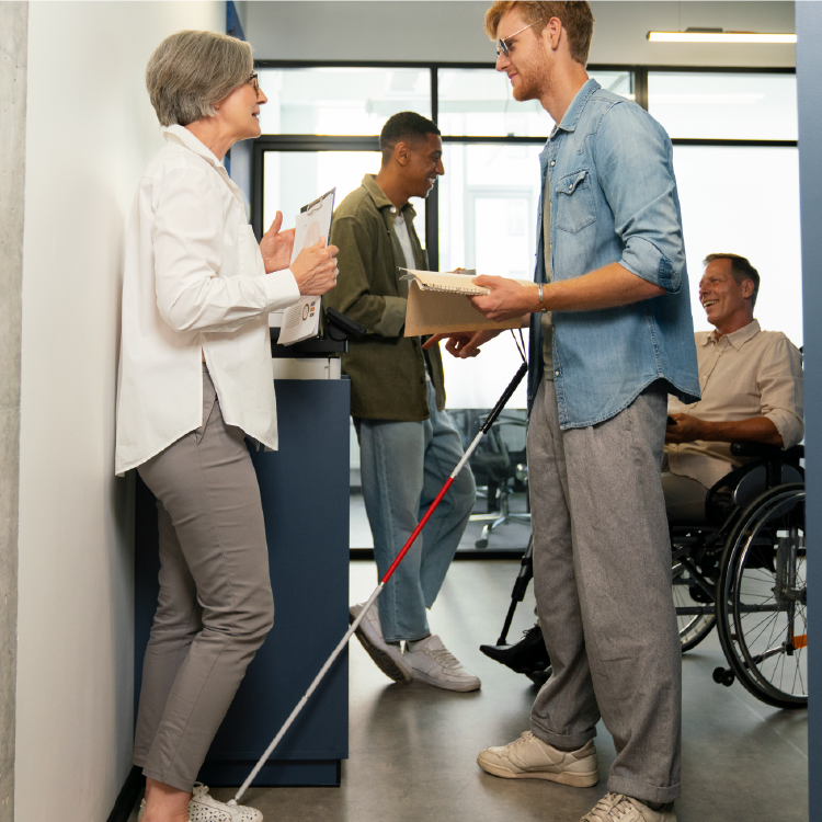 משרד, ובו אדם עיוור מחזיק מקל ומחברת ברייל מדבר עם עובדת מבוגרת, מאחוריהם עובד על כיסא גלגלים מדבר עם עובד קהה אור.