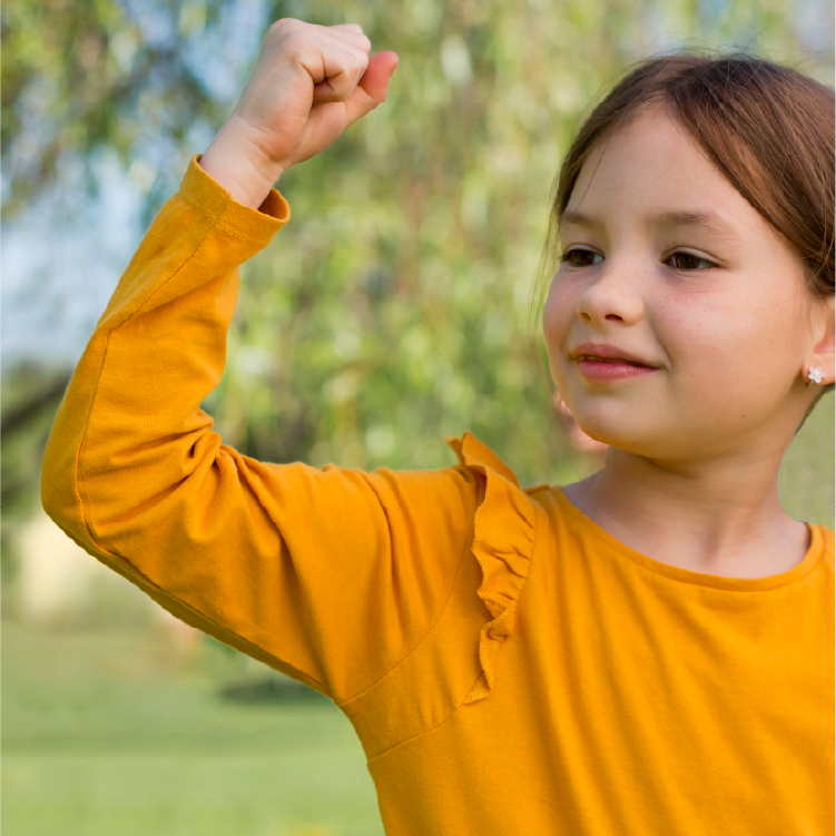 ילדה בהירה עם חולצה צהובה על רקע של טבע מטושטש עושה תנועה של שריר ביד - כוח